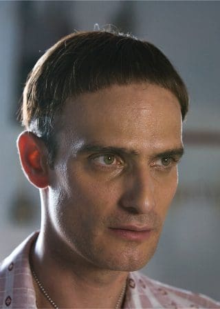 A close-up of a man with a distinctive bowl haircut and serious expression. He has light green eyes, a neutral skin tone, and is wearing a patterned shirt. The background is softly blurred.