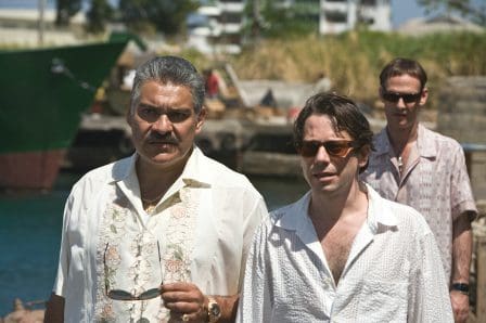 Three men walking along a dock on a sunny day. The first man, wearing a white shirt, holds sunglasses and has a serious expression. The second man, in a white shirt, looks slightly apprehensive. A third man in sunglasses follows behind, wearing a patterned shirt.