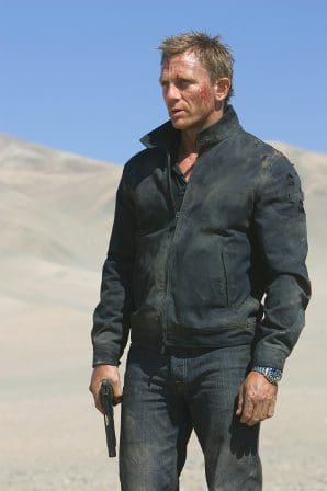 A man standing in a desolate landscape, wearing a black leather jacket and jeans, holding a gun. His face shows signs of dirt and wear, contrasting against the clear blue sky and distant mountains.