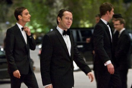 A group of men in black tuxedos walking outside at night. One man is smiling confidently as they approach a luxury vehicle. Soft lights from surrounding trees create an elegant ambiance.