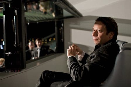 A man in formal attire sits pensively on a sleek gray couch, looking out through a large window. In the background, blurred figures in tuxedos engage in conversation, suggesting an elegant event.