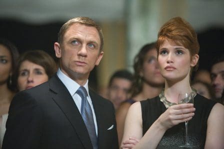 A well-dressed man in a tuxedo and a woman in an elegant black dress stand together, both looking intently at something in the distance. They appear focused amid a crowd of elegantly dressed people at a sophisticated event.