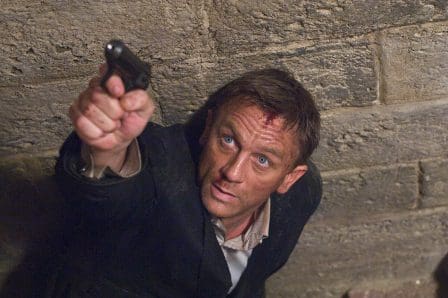A man with tousled hair and blue eyes, gripping a handgun while looking up with a determined expression in a dimly lit, stone-walled environment.