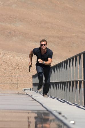 A man wearing sunglasses runs on a narrow metallic structure with a desert landscape in the background. He holds a handgun and wears a fitted black shirt and jeans, demonstrating urgency and focus.