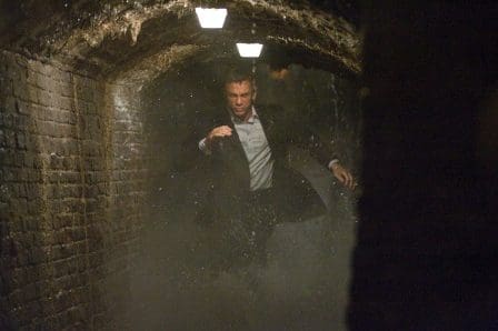 A man in a suit running through a dimly lit underground tunnel, with water splashing around him. The walls are made of brick, and moisture is visible in the air, creating a tense atmosphere.
