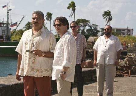 A group of four men standing on a dock by the water, with boats in the background. The men are dressed in light-colored shirts and relaxed attire, suggesting a warm climate. Palm trees sway in the breeze under a partly cloudy sky.