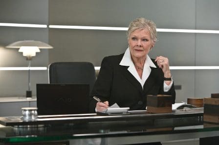 A mature woman with short gray hair sitting at a modern office desk. She wears a black blazer with a white shirt, holding a pen and looking thoughtfully at a notebook, with a laptop and wooden blocks on the desk.
