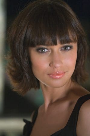 A woman with dark, shoulder-length hair and bangs smiles softly at the camera. She has a natural makeup look enhancing her features and a subtle gloss on her lips. The background is softly blurred, emphasizing her presence.