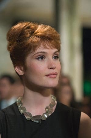 A woman with stylish short red hair and subtle makeup, wearing a black dress and a chunky necklace, gazes thoughtfully in a softly lit event venue, with blurred figures in the background.