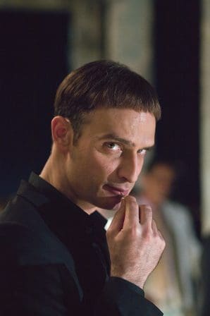 A man in a black suit with short hair looks directly at the camera, partially biting his finger, in a softly lit setting. Out-of-focus figures are visible in the background, suggesting an intimate gathering.