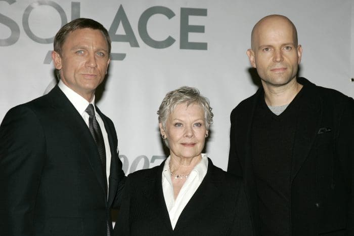 Three individuals pose for a photo at a promotional event. The first person on the left is dressed in a black suit and tie, the second person in the center wears a black jacket with white blouse and has short hair, while the third person on the right is in a dark sweater.