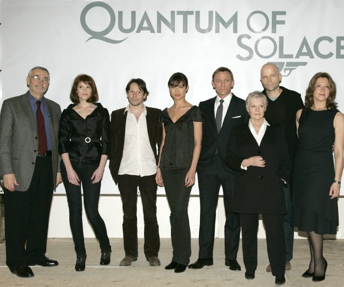 A group of eight individuals poses in front of a promotional banner for "Quantum of Solace." They are dressed in formal attire, showcasing a variety of styles. The setting is a well-lit event space, emphasizing the film's theme.