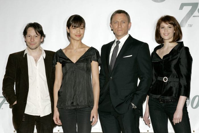 Four individuals posing together at a promotional event, dressed in formal attire. The group includes two men and two women, with varying styles, set against a light background featuring promotional text.