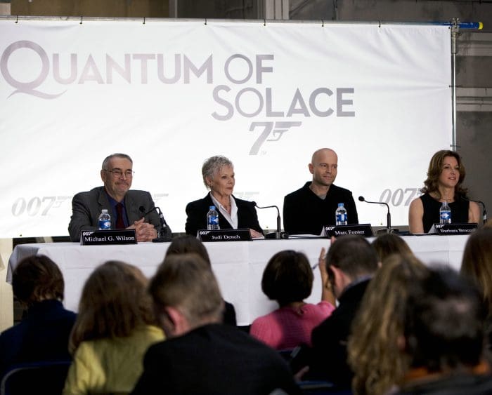 A panel of four individuals at a press conference for "Quantum of Solace," with a large banner in the background. The panelists include a man in a dark suit, a woman with short gray hair, a man in a black suit, and a woman with long hair, all seated at a table with microphones.