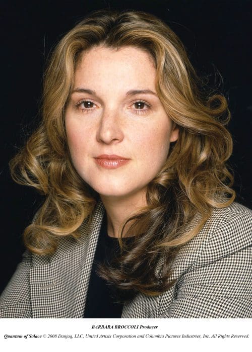A woman with wavy, shoulder-length hair and a neutral expression, wearing a black top under a checkered blazer, set against a dark background. She appears poised and professional, with subtle makeup enhancing her features.