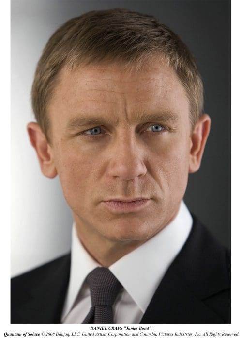Close-up portrait of a man with light brown hair and blue eyes, wearing a dark suit with a white shirt and a tie. He has a serious expression, highlighting his strong features against a softly blurred background.