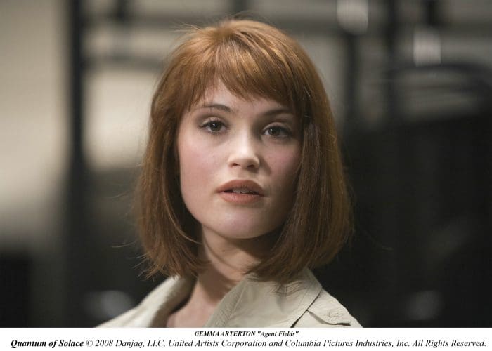 A woman with short brown hair and a neutral expression, wearing a light-colored jacket, is looking directly at the camera. Soft lighting highlights her features against a blurred background, suggesting an indoor setting.