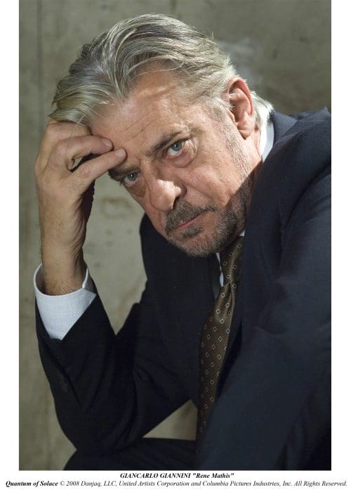A middle-aged man in formal attire rests his chin on his hand with a contemplative expression. He has grey hair, and the setting features a textured wall, suggesting a somber atmosphere.