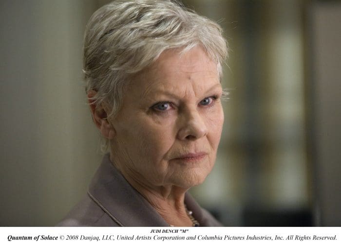 A close-up portrait of an older woman with short, silver hair, looking intently with a serious expression. She is dressed in a gray blazer, with a blurred background, conveying a sense of focus and determination.