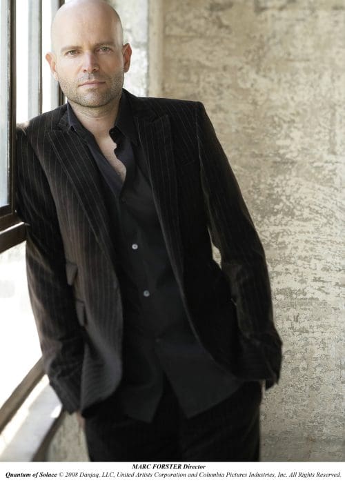 A man in a pinstripe suit leans against a window in an industrial-looking space. His gaze is focused, and he appears confident, with a bald head and slight smile. Natural light illuminates the textured wall behind him.