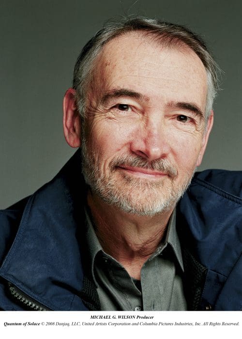 A senior man with a silver beard and short hair smiles warmly at the camera. He is wearing a dark jacket over a light shirt, set against a soft, neutral background that adds a professional feel to the portrait.