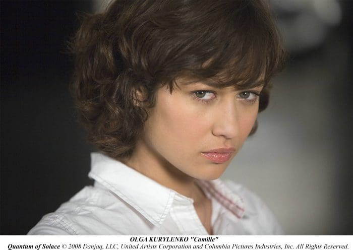 A woman with short, curly hair looks intensely at the camera, wearing a white blouse. The background is softly blurred, emphasizing her facial expression and features.