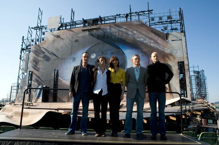 Five individuals standing in front of a large stage with a dramatic backdrop. The stage features an intricate design that resembles an eye, and the people are dressed in formal attire, posing for a photo under clear skies.