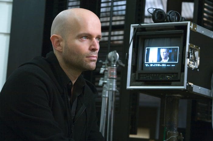 A man with a shaved head sitting pensively beside a video monitor, watching a scene being filmed. He is dressed in a black hoodie, with studio equipment and a soft focus background.
