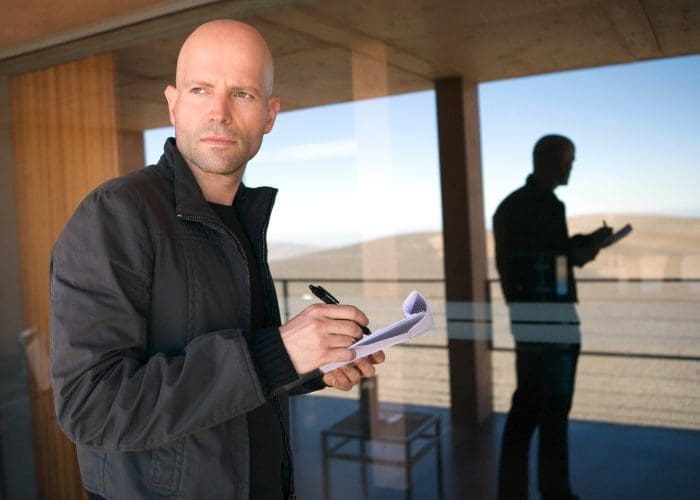 A bald man in a black jacket stands outdoors, writing notes in a notepad. He gazes thoughtfully at the horizon, with a reflected silhouette of himself visible in a glass wall behind him.