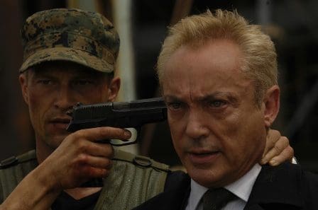 A tense scene featuring a soldier holding a gun to the head of a man in a suit. The background is blurred, focusing on their expressions. The soldier wears a camouflage cap, while the man appears anxious and serious.
