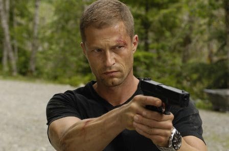 A man with short blond hair and a serious expression holds a handgun in a forested area. He has visible bruises and scrapes on his face, indicating a recent conflict. The background features tall trees and a gravel path.