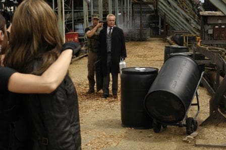 A man in a long coat stands beside a worker in a warehouse-like setting. Two large black barrels are positioned nearby, and part of a group of people is shown in the foreground, partially obscured.