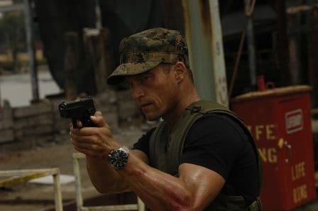 A muscular man in military attire holds a handgun, aiming it with focus. He wears a camouflage cap and an olive green vest, with a watch visible on his left wrist, set against an industrial backdrop.