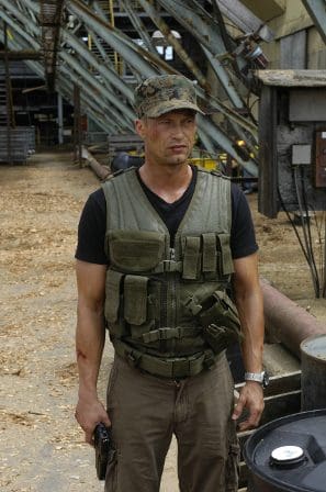 A man wearing a tactical vest and camouflage cap stands in an industrial setting, surrounded by wooden debris and metal structures, holding a gun at his side, with a serious expression.