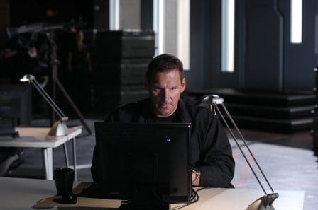 A man with a serious expression sits at a desk in a dimly lit room, focused on a computer screen. The workspace features minimal decor, with a desk lamp and a black mug beside him, emphasizing a tense atmosphere.