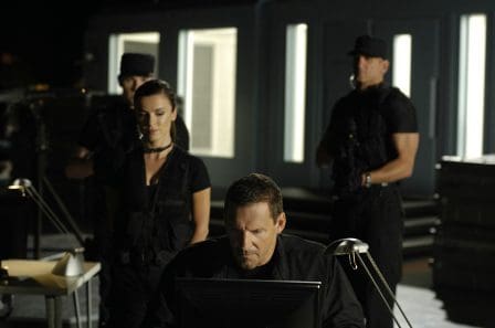 A team of four individuals dressed in tactical black clothing stands in a dimly lit room. The focus is on a man sitting at a desk, intently looking at a computer screen. Two armed colleagues stand behind him, while a woman watches closely.