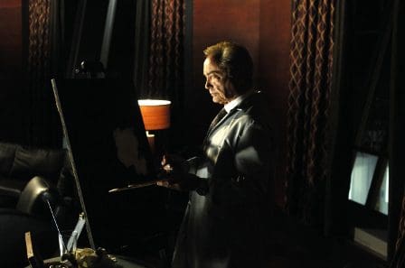 An artist in a white coat is focused on painting at an easel in a dimly lit room. A warm lamp illuminates the space, highlighting his thoughtful expression and the textures of the painting.