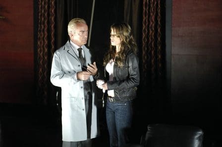 A man in a white lab coat talks to a woman wearing a black leather jacket and jeans, both standing in a dimly lit room with dark walls and rich curtains.