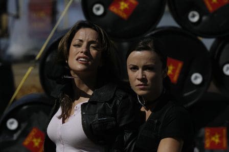 Two women wearing tactical vests stand confidently in front of large, black barrels marked with symbols. They have determined expressions and their hair is styled casually, blending an adventurous spirit with a sense of readiness for action.