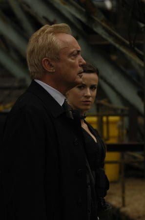 A man with light blond hair in a black coat stands in profile, looking attentively into the distance, next to a woman in a black outfit. The background features industrial elements and yellow containers.