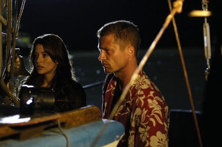 A man and a woman standing on a boat at night, with the woman wearing a black top and the man wearing a colorful floral shirt. They appear focused and engaged in conversation, with a blurred water background illuminated by soft light.