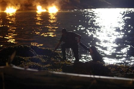 A person crouching near rocky terrain at night, while another person is partially submerged in water. The shimmering surface of the water reflects distant flames on the shore, creating a dramatic contrast with the dark surroundings.