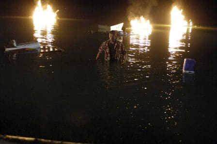 A person standing in shallow water at night, surrounded by floating debris. Flames erupt from several points on the water's surface, creating a dramatic contrast against the dark surroundings.