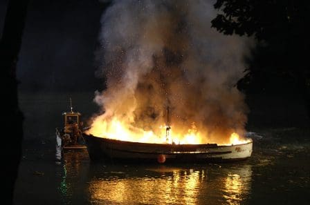 A fishing boat engulfed in flames, illuminated by bright orange fire, while a small vessel approaches nearby, creating ripples on the water's surface at night.
