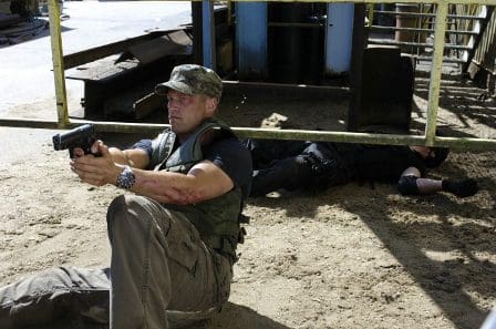A man in tactical gear sits on the ground, aiming a handgun in front of him. The sandy terrain is scattered with debris, and a figure in dark clothing lies motionless in the background, partially obscured by a metal structure.