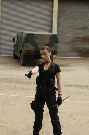 A woman dressed in tactical black clothing, holding a handgun confidently while pointing it forward. She wears tactical gear, including a vest, gloves, and a utility belt. In the background, a camouflaged military vehicle is parked near a closed metal garage.