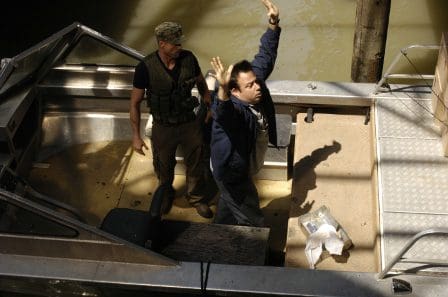 A man with raised hands stands on a boat, appearing to surrender. Behind him, a man in tactical gear watches attentively. The scene is set on a riverbank, with sunlight illuminating the surroundings.