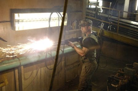 A soldier wearing military gear operates a welding tool inside an industrial setting, sparks flying as they work on a metal pipe. The environment is dimly lit and filled with machinery.