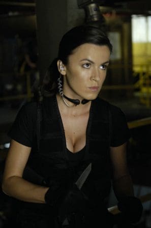 A woman in tactical attire, wearing a headset and holding a knife, stands alert in an industrial setting. Her expression is focused, and the background features dim lighting and partial views of machinery.