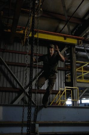 A person in work attire performing an action on a steel beam in a warehouse, surrounded by industrial structures and chains, with yellow safety rails visible in the background.